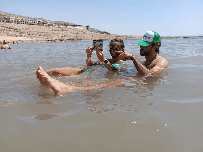Young travelers floating in the Dead Sea in Jordan, wearing mineral-rich mud masks and laughing together.