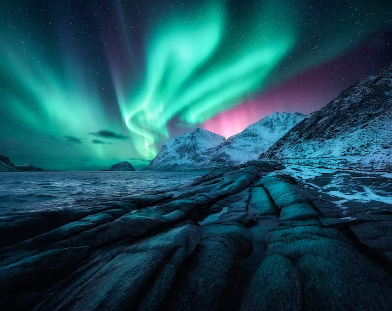 Green aurora borealis swirling above a rocky coastline and snowy peaks, an affordable northern lights destination