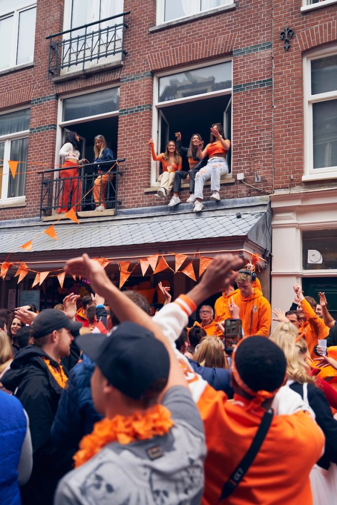 People sitting in open windows watching and celebrating King’s Day on a busy Amsterdam street.