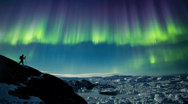 Photographer capturing the Northern Lights over an icy landscape in Greenland under a glowing green aurora