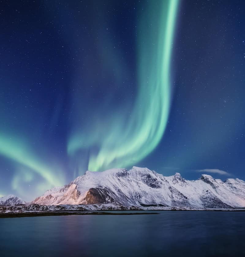Bright green aurora borealis over calm water and snow-covered mountains in Iceland, a budget-friendly northern lights spot