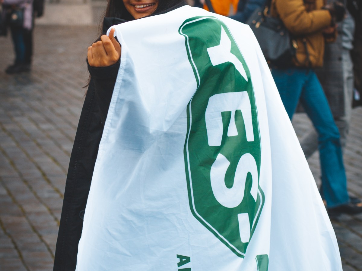 Person wearing a green cap and holding a large 'YES-TRIP' banner in a cobblestone plaza.