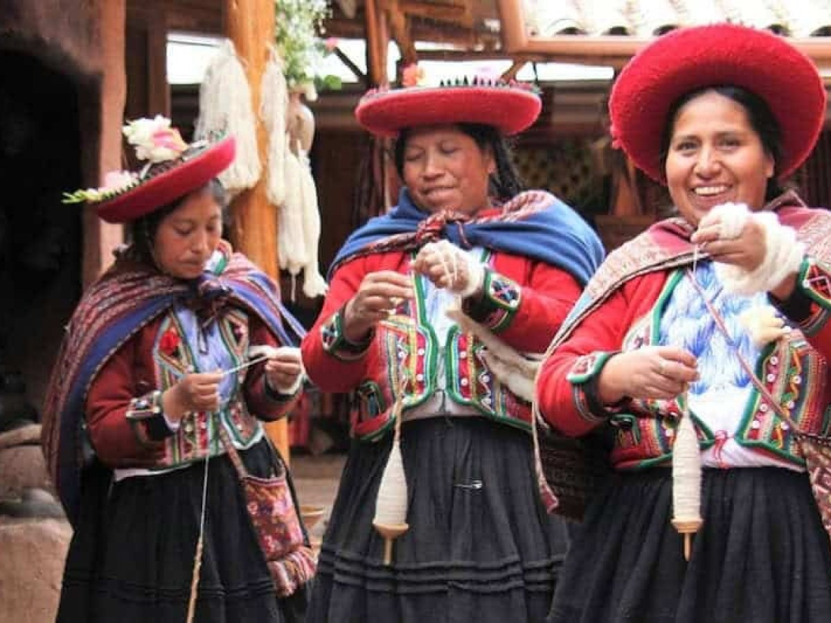 Three women in traditional attire spinning yarn with spindles, smiling and standing indoors.