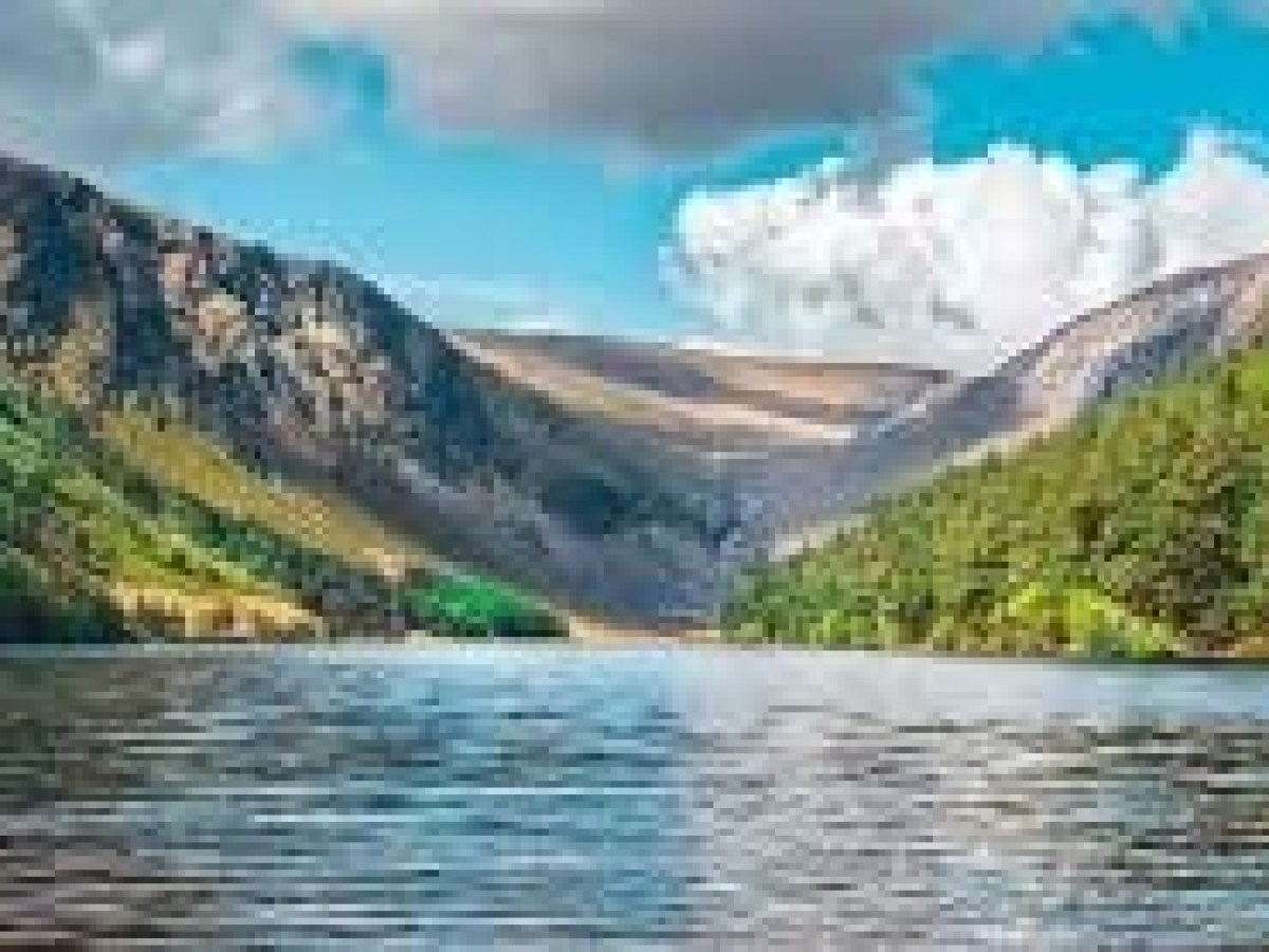 Scenic lake surrounded by green forests and mountains under a blue sky with clouds.