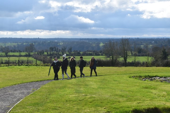 Five people walking on a path in a green field under a cloudy sky.