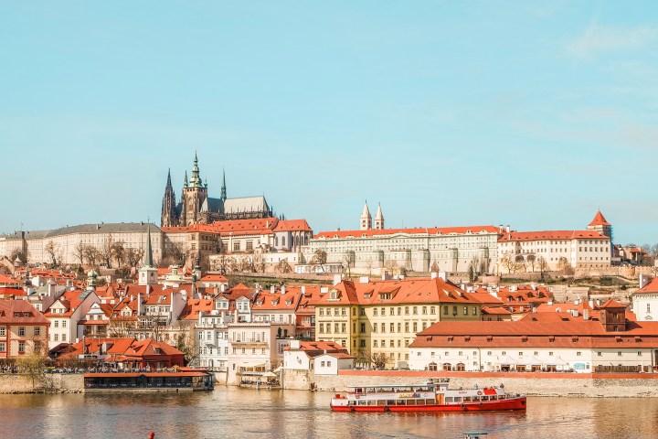 Scenic view of a historic city with a castle on a hill, red-roofed buildings, and a river with a boat.