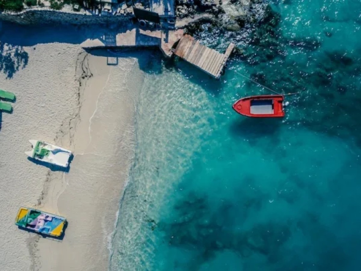 Aerial view of sandy beach with sun loungers and a red boat on turquoise water.