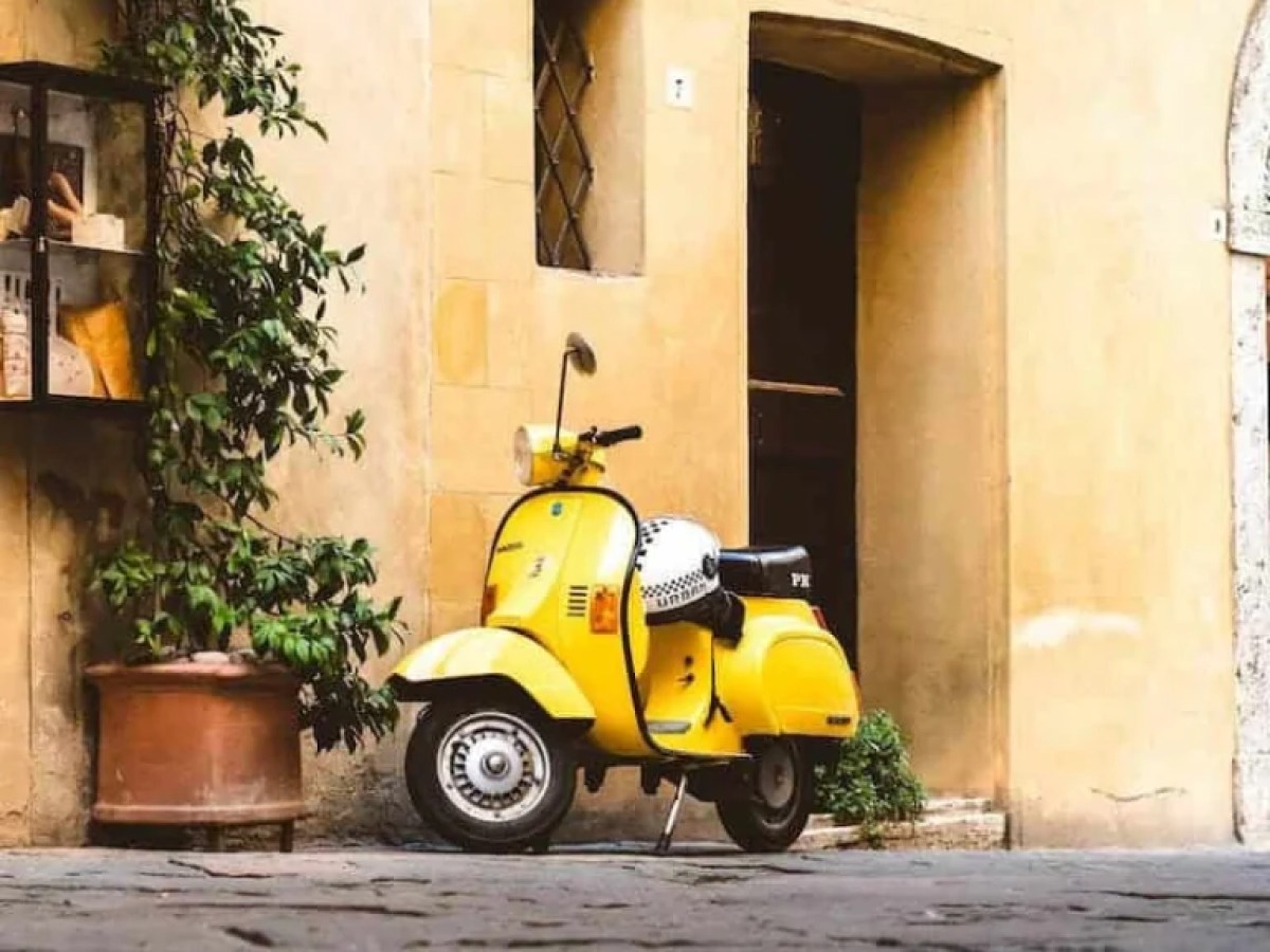 Yellow scooter parked on a cobblestone street by a beige wall with a window and potted plant.
