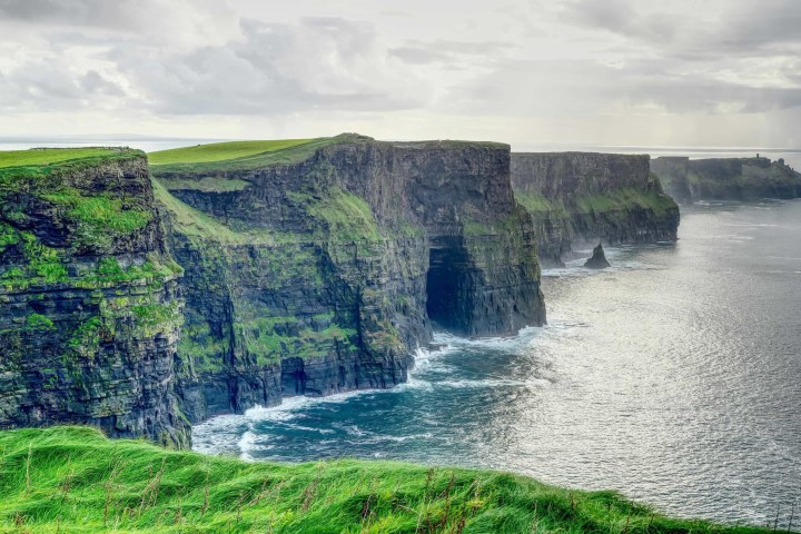 Dramatic cliffs along a coastline with green grass and a cloudy sky.