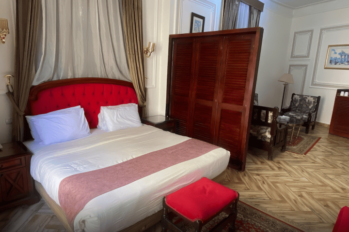 Hotel room with a bed, red headboard, armchairs, and wooden screen on a patterned floor.