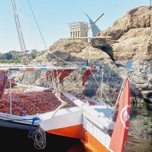 Boat docked at rocky shore with windmill and historical building in the background.
