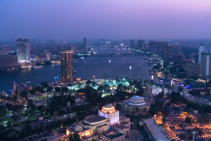 Aerial view of a cityscape at dusk with a river and illuminated buildings.