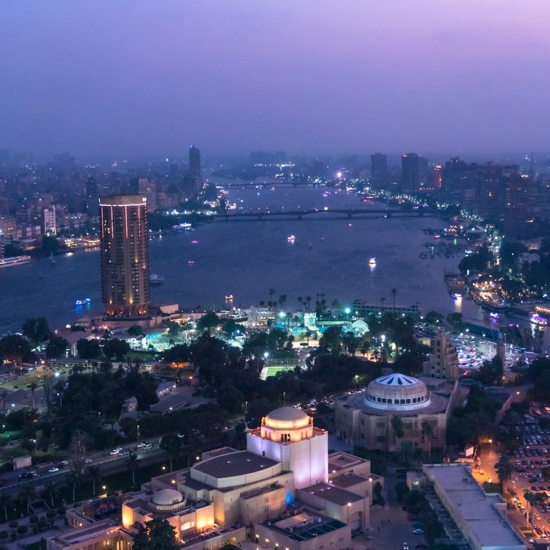 Aerial view of a cityscape at dusk with a river and illuminated buildings.