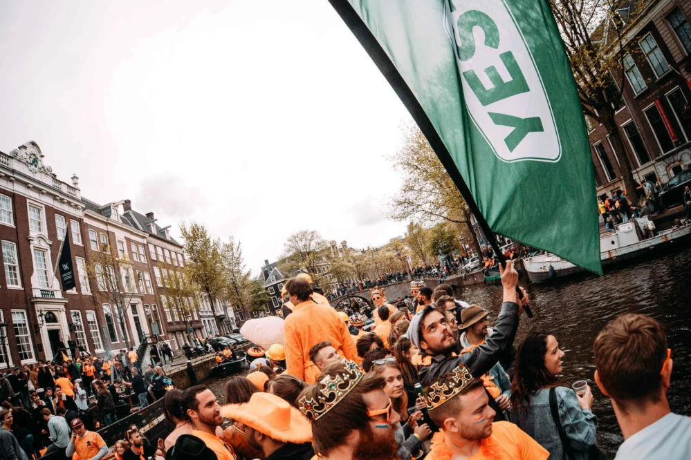 Crowd in orange outfits celebrating by a canal, man holding a 'YES' flag.