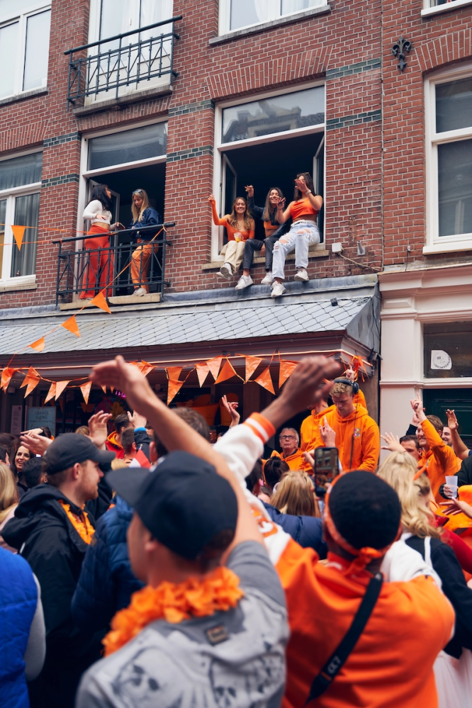 People in orange celebrate on a street and balcony during a festive event.