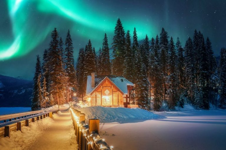 Snowy cabin in forest with northern lights and lit path at night.