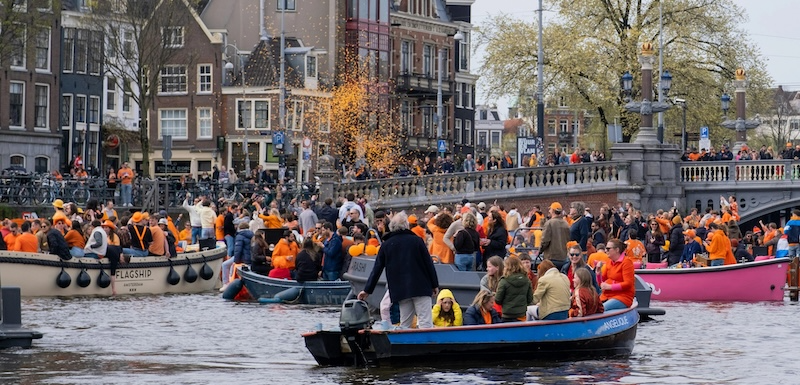 Crowd in boats on a canal, many wearing orange, with historic buildings in the background.