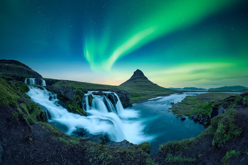 Waterfall and mountain under a vibrant aurora borealis in a night sky.