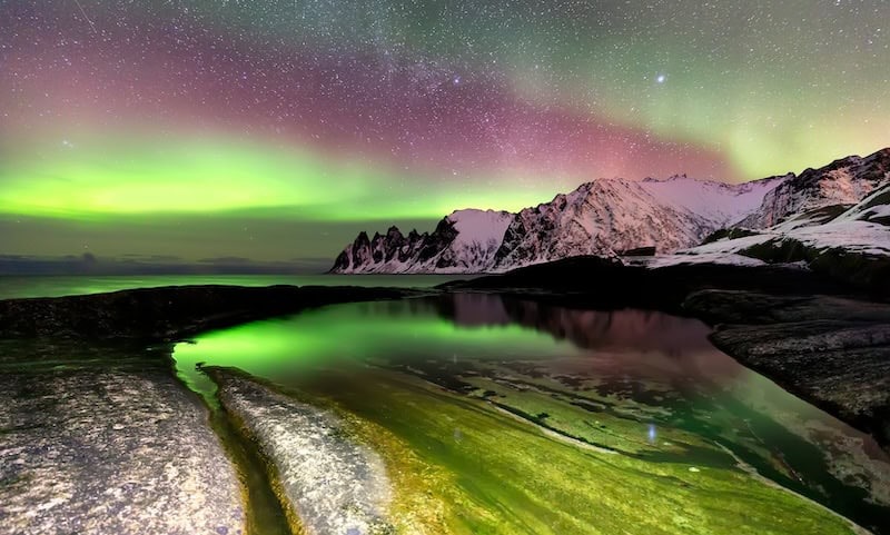Aurora borealis over snowy mountains and reflective water at night.